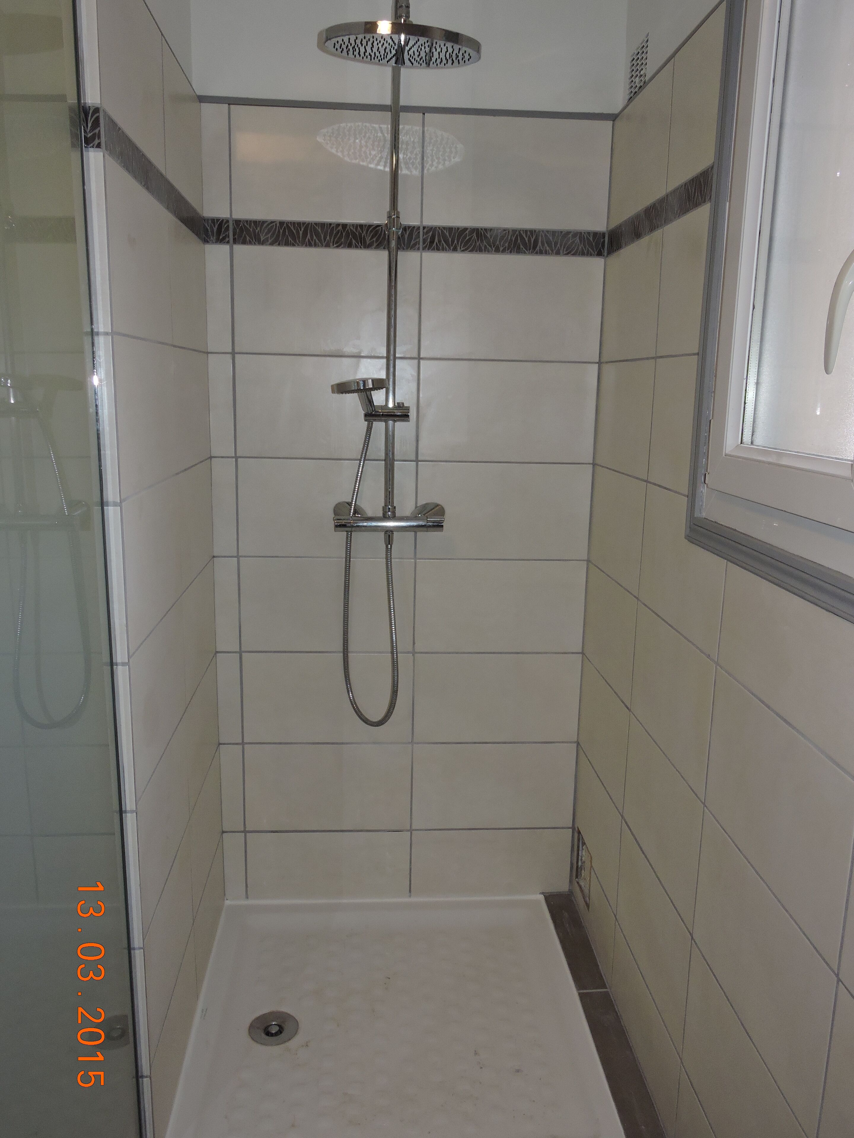 Une douche équipée d’un pommeau de douche à main et d’un pommeau de douche fixe au plafond, d’une porte en verre et d’un sol carrelé.