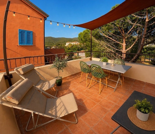 A terrace with a table and chairs, a potted plant, and a building with blue shutters.