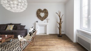A living room with a dark brown sofa, a patterned rug, a white fireplace, and a large window. - L’Écume-Maison confortable et équipée (Saint-Coulomb)