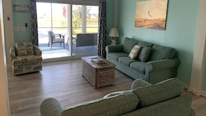 A living room with a green couch, a wooden coffee table, and a chair with a beach-themed cushion. There is a painting on the wall and a window with curtains. - Salty dog on the rocks (Port St Joe)