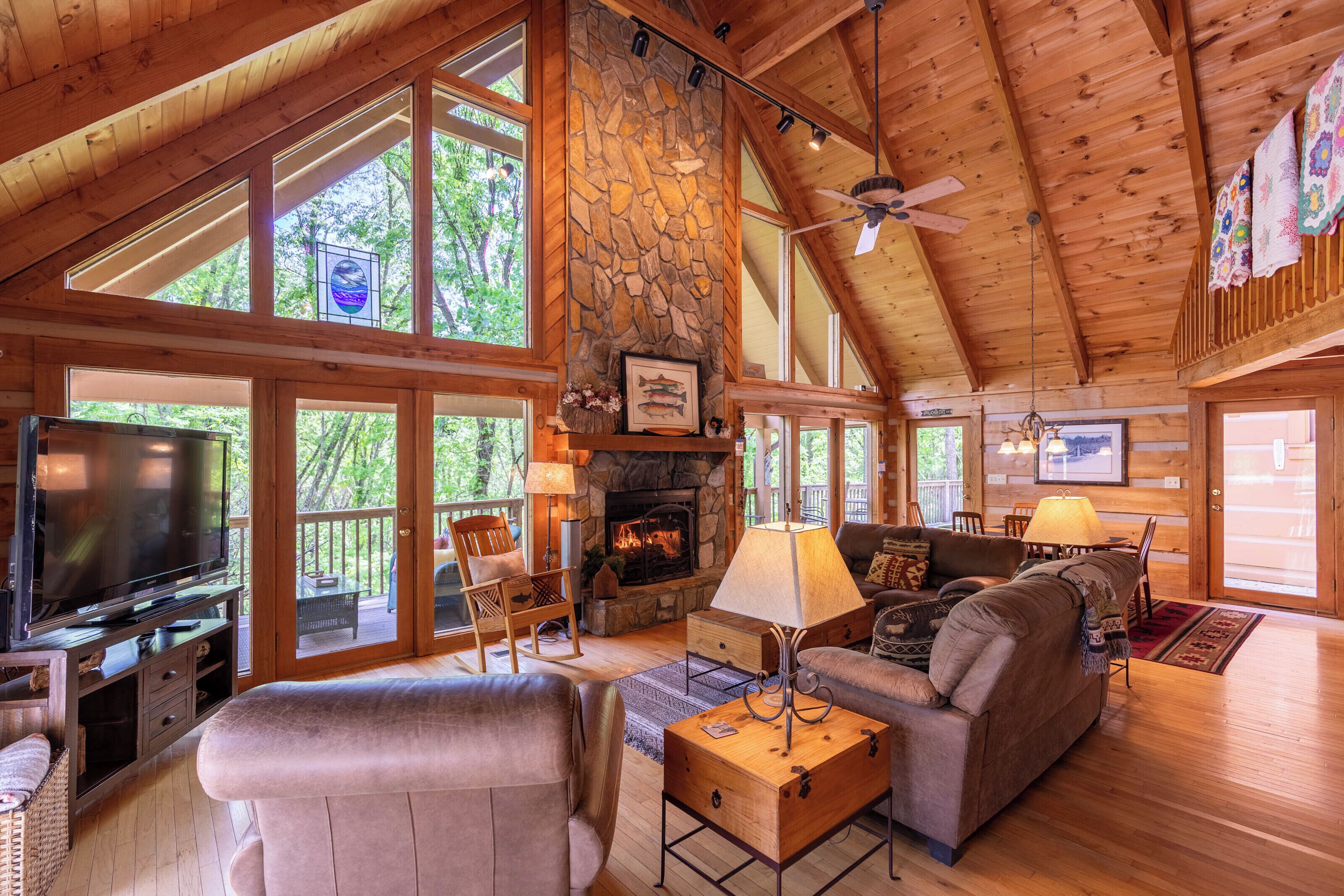 A spacious cabin interior with a stone fireplace, wooden furniture, and large windows.