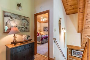 Un pasillo con techo de madera, un cuadro enmarcado, una lámpara y una cama visible a través de una puerta abierta.