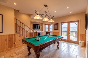 Une table de billard dans une pièce aux murs lambrés, avec un escalier et une vue dégagée sur l’extérieur grâce à de grandes fenêtres.