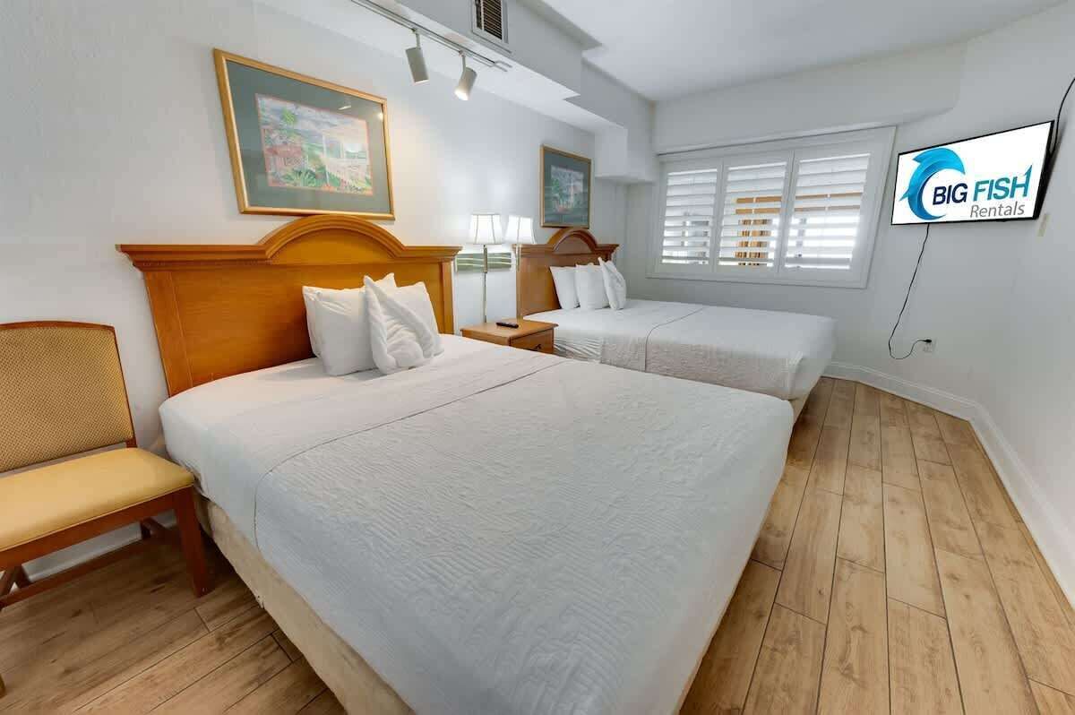 A hotel room with two beds, a wooden headboard, a bench, a TV, and a window with blinds.