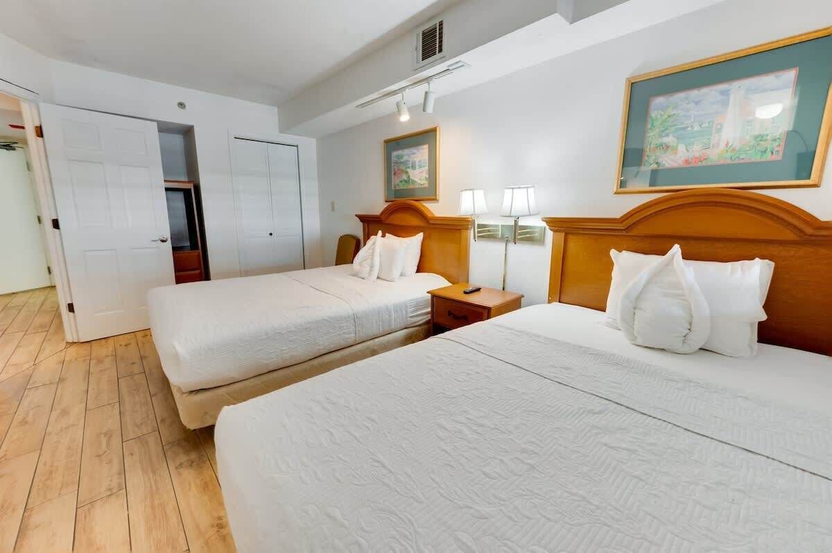 Habitación de hotel con dos camas, cabecero de madera, mesita de noche, lámpara y un cuadro en la pared.