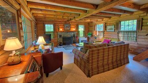 Interior de una cabaña de troncos con chimenea, muebles de madera y ventanas amplias.