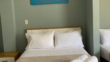 Una cama bien tendida con sábanas blancas y una toalla doblada, cabecera de madera y un letrero en la pared con coordenadas.