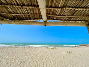 A beach with a thatched roof structure, sandy beach, and ocean view. - Castillo del Mar Suites (Riohacha)