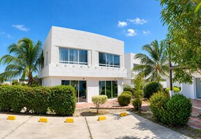 Un edificio moderno de dos pisos con fachada blanca, ventanas amplias y un área de entrada pavimentada con pilares amarillos.