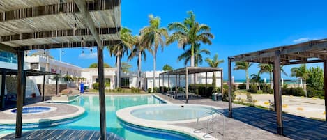 Un área de piscina con varias piscinas circulares, palmeras y una zona de estar cubierta.