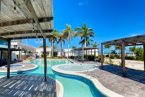 Un área de piscina con varias piscinas circulares, palmeras y una zona de estar cubierta.