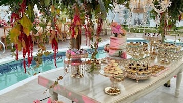 Un espacio interior elegante con una larga mesa que exhibe una variedad de postres y una piscina al fondo.