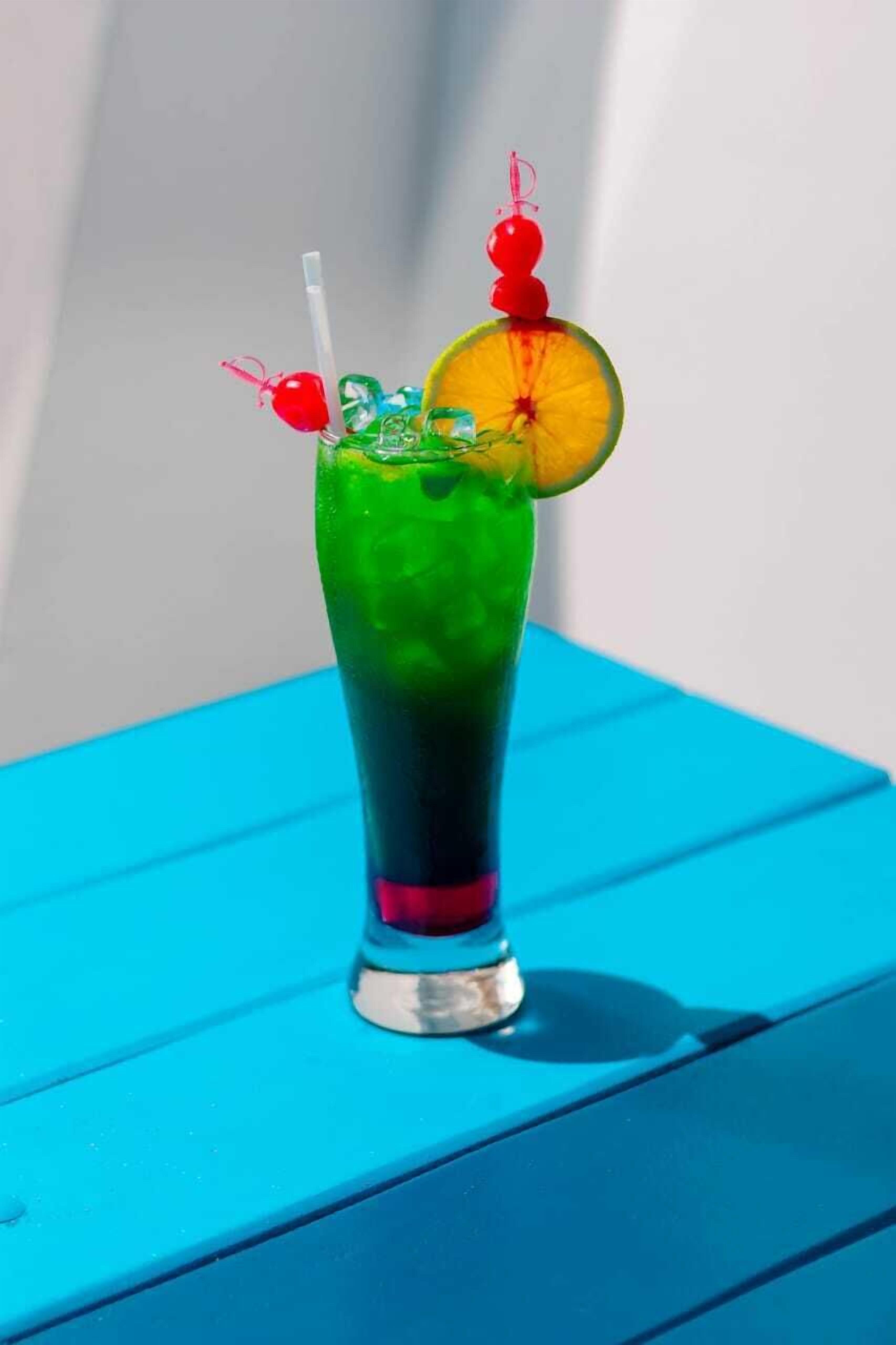 A tall glass with a gradient of green to blue, containing a slice of orange and a red cherry on a skewer, placed on a blue surface.