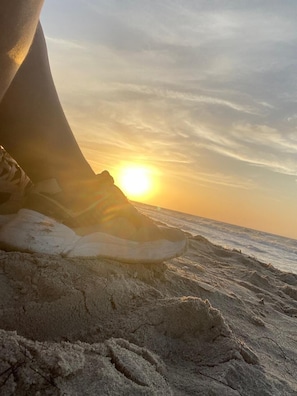 A beach scene with a sunset, sand, and a person's foot in a sneaker.