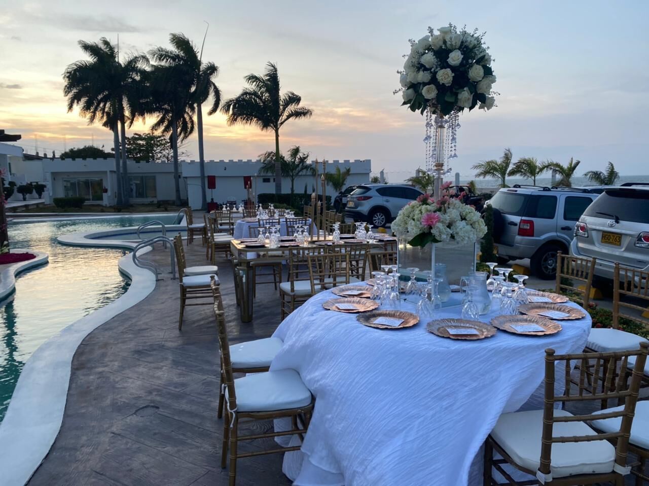 Un espacio exterior con mesas listo para una comida, con vistas a una piscina y palmeras.