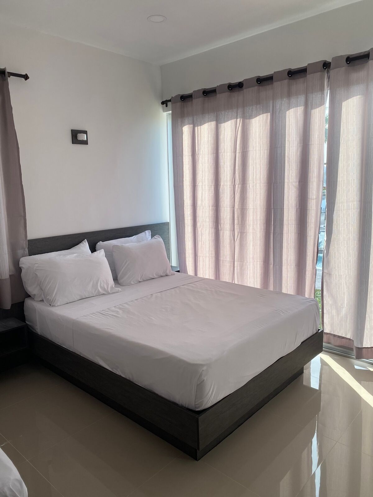 Una habitación de hotel moderna con una cama grande, cabecera oscura y cortinas de gasa.