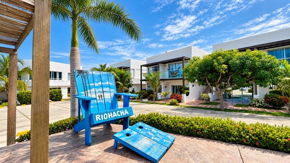 Una silla azul con el texto 'RIOHACHA', un edificio moderno y un jardín cuidado.