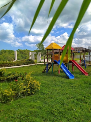 A playground with slides and a small shelter, surrounded by grass and trees.