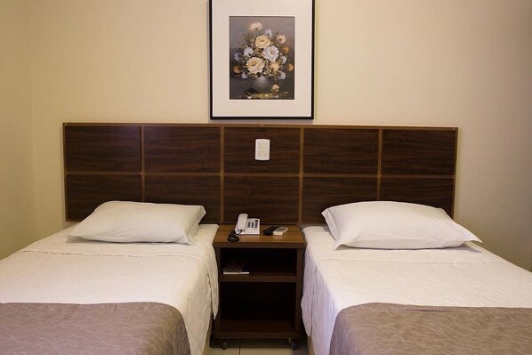 Habitación de hotel con dos camas individuales, cabecera de madera, mesita de noche con teléfono y un libro, y una obra de arte floral enmarcada en la pared.