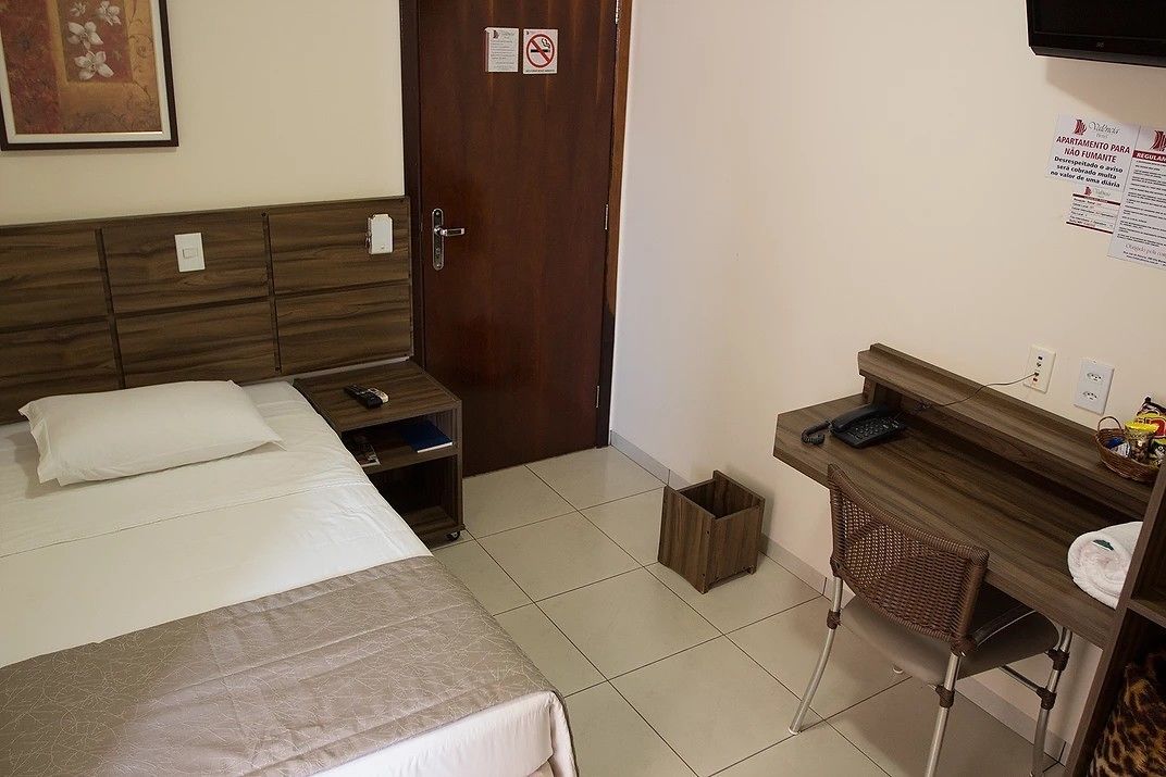 A hotel room with a bed, a desk with a chair, a TV, and a door with a sign.