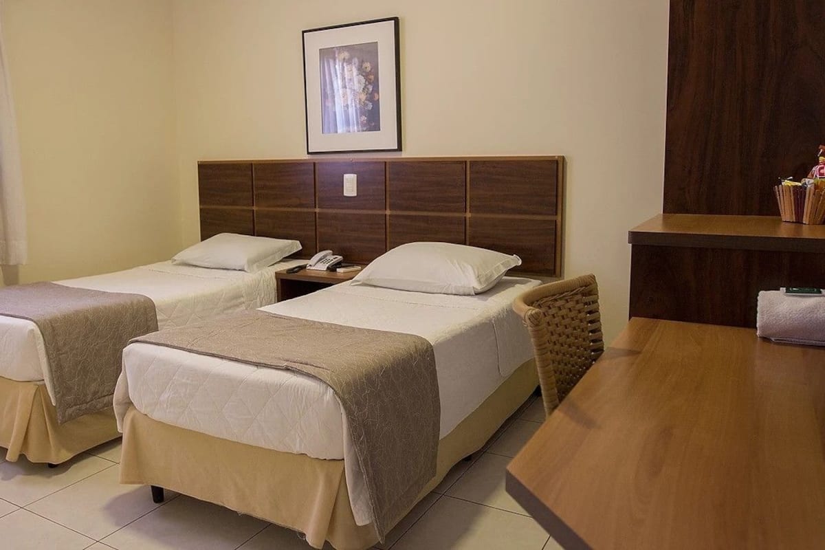 A hotel room with two beds, a wooden headboard, a bedside table with a lamp, and a framed picture on the wall.