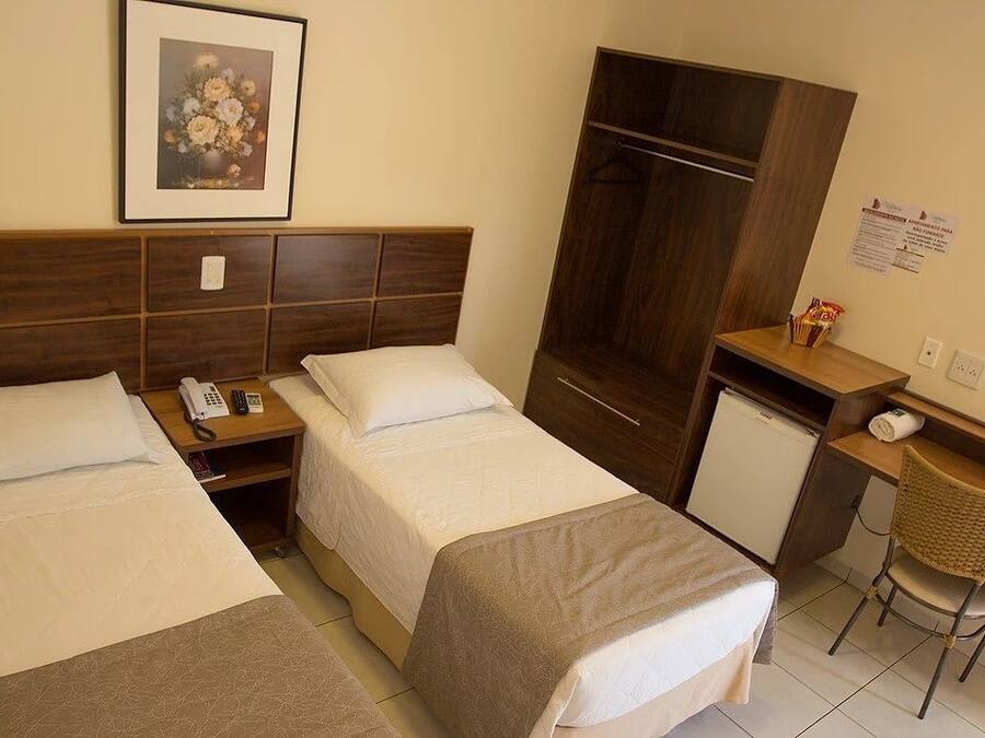 A hotel room with two beds, a desk, a chair, a wardrobe, and a small refrigerator.