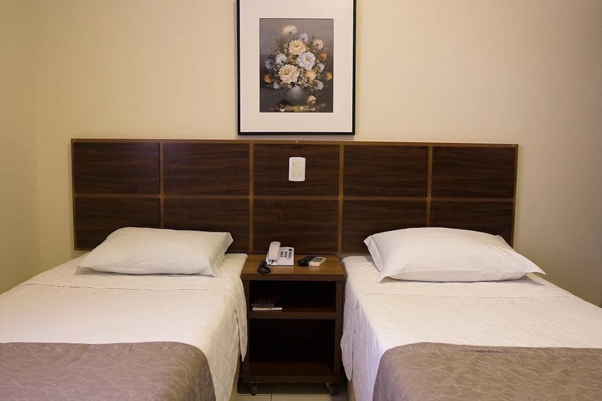 A hotel room with two single beds, a wooden headboard, a bedside table with a phone and a book, and a framed floral artwork on the wall.