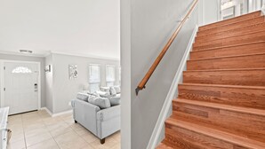 A staircase with wooden steps and a handrail, leading to a landing with a white door and a gray couch in a room with light-colored walls and a tiled floor.