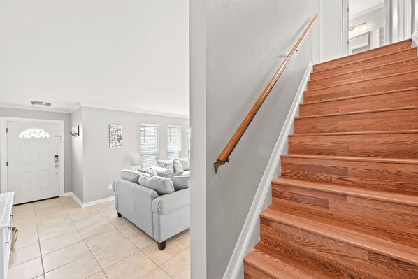 A staircase with wooden steps and a handrail, leading to a landing with a white door and a gray couch in a room with light-colored walls and a tiled floor.