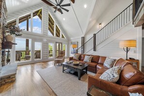 A spacious living room with a high ceiling, a large sofa set, a coffee table, and a staircase leading to an upper level.