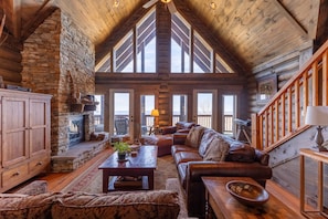 A log cabin interior with a stone fireplace, leather sofa, wooden coffee table, and a staircase. - Highland Mist  Pet Friendly, Beautiful Views, Hot Tub, Game Tables, Winery Close by (Banner Elk)