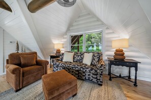 Une chambre à coucher confortable, aménagée en mezzanine, avec un plafond en pente, un canapé, un fauteuil, une petite table et une fenêtre donnant sur un paysage verdoyant.