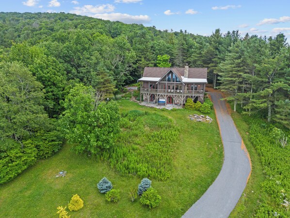 A large, two-story house with a wrap-around porch, surrounded by dense forest and a winding road.