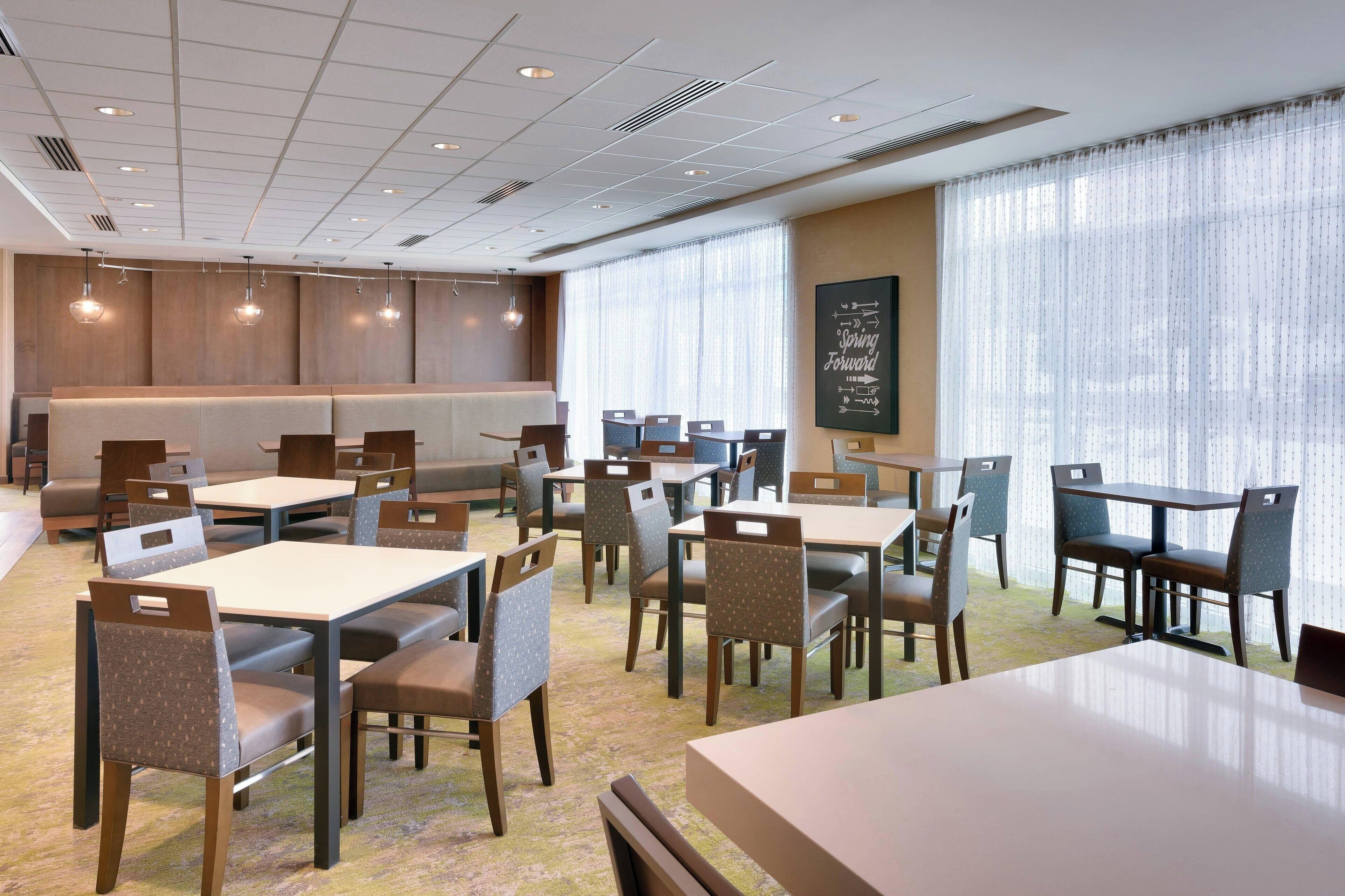 A modern dining area with multiple tables and chairs, a large window with sheer curtains, and a motivational quote on the wall.