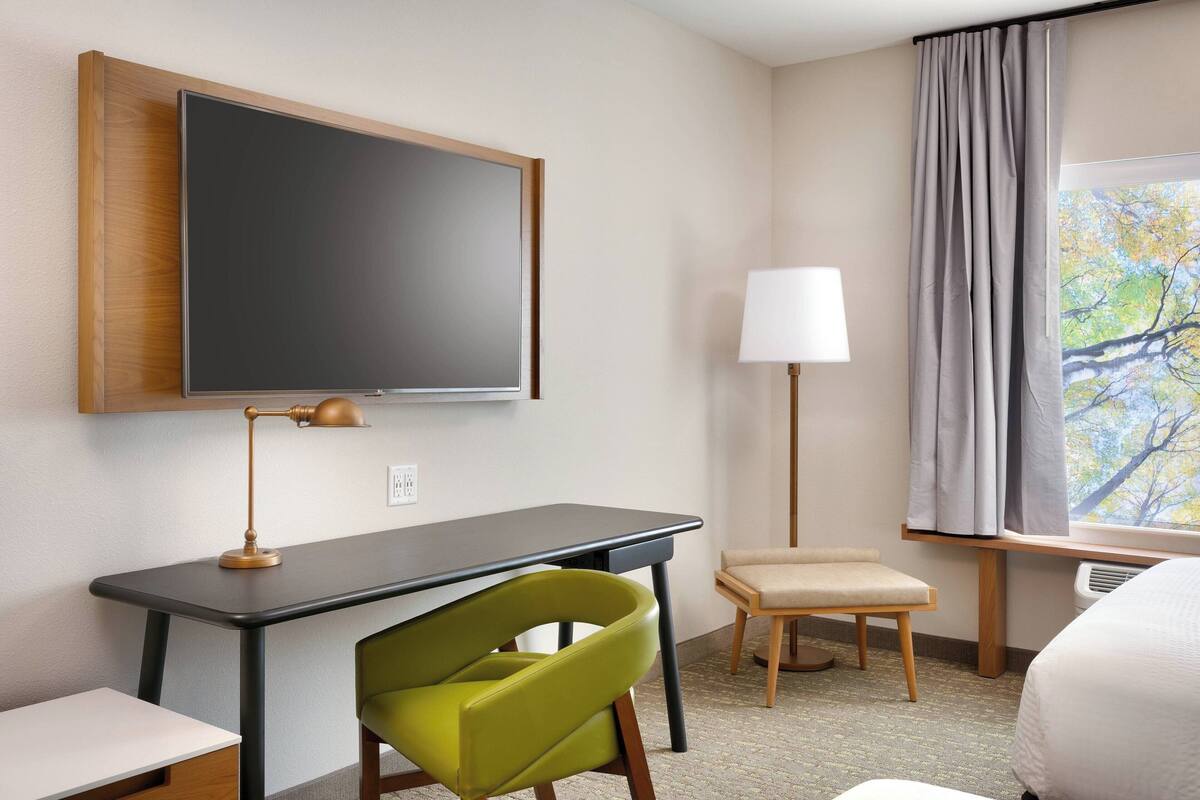 A hotel room with a desk, a green chair, a bed, a TV, a lamp, and a window with curtains.