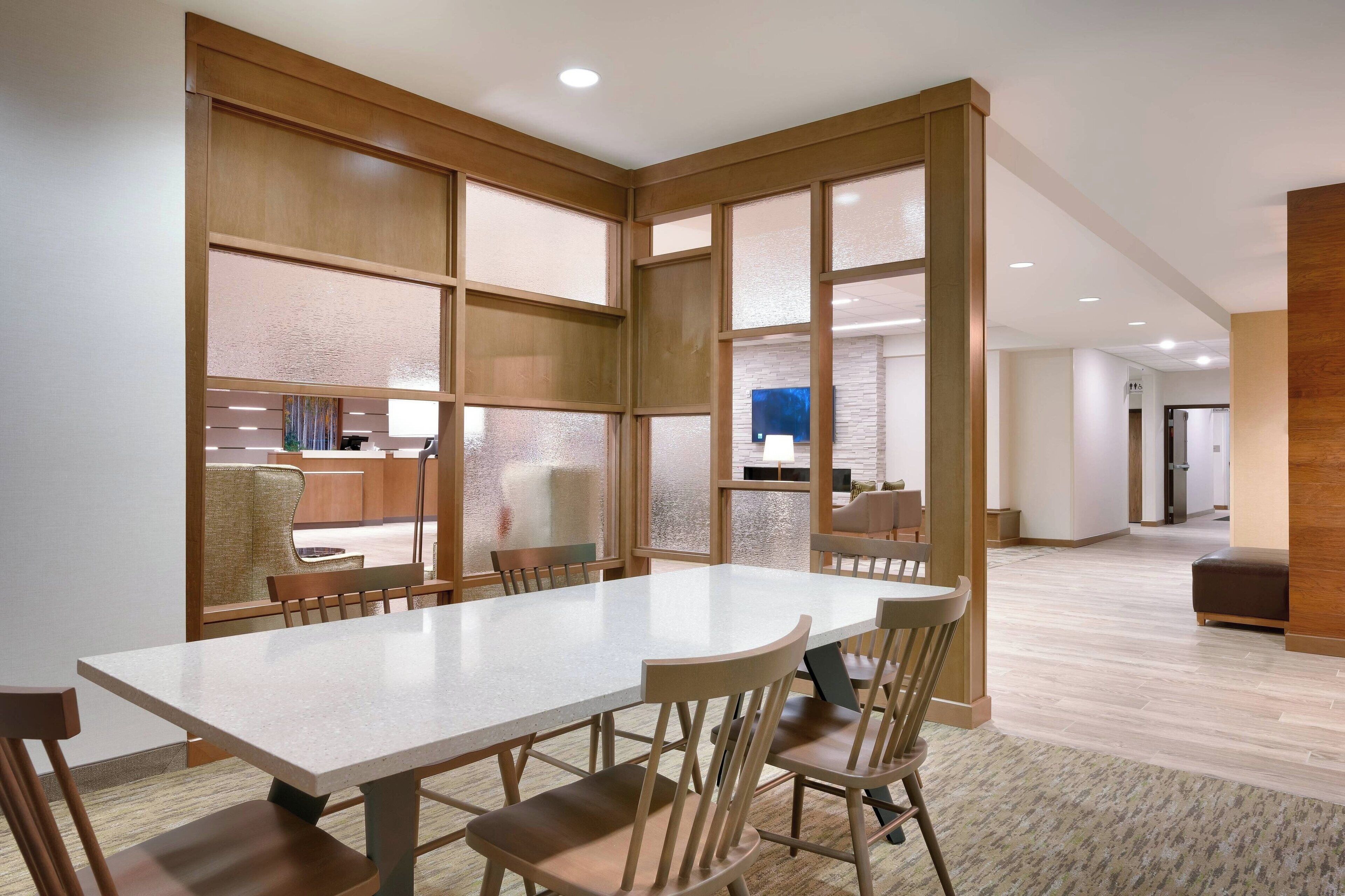 A modern dining area with a long table and chairs, wooden partitions, and a view of the kitchen in the background.