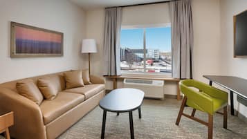 A hotel room with a beige sofa, a small white table, a green chair, a large window with curtains, and a painting on the wall.