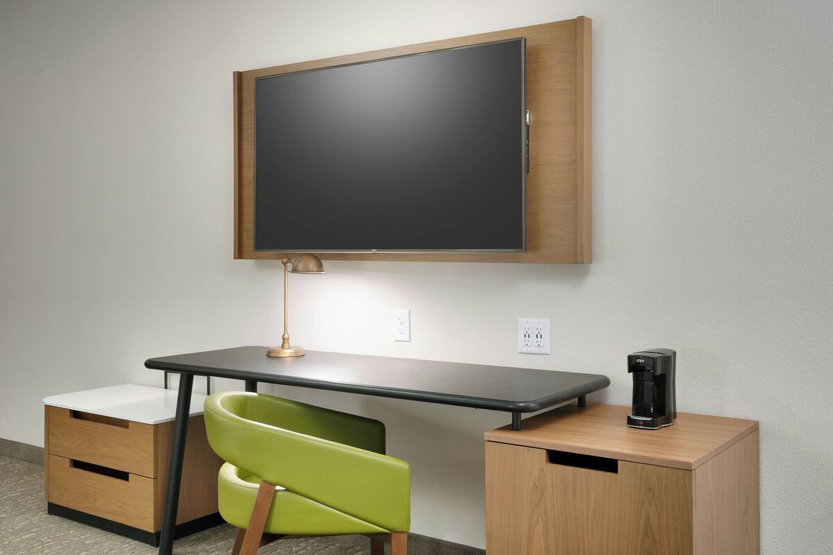 A modern office desk with a flat-screen monitor, a desk lamp, a coffee maker, and a green chair.