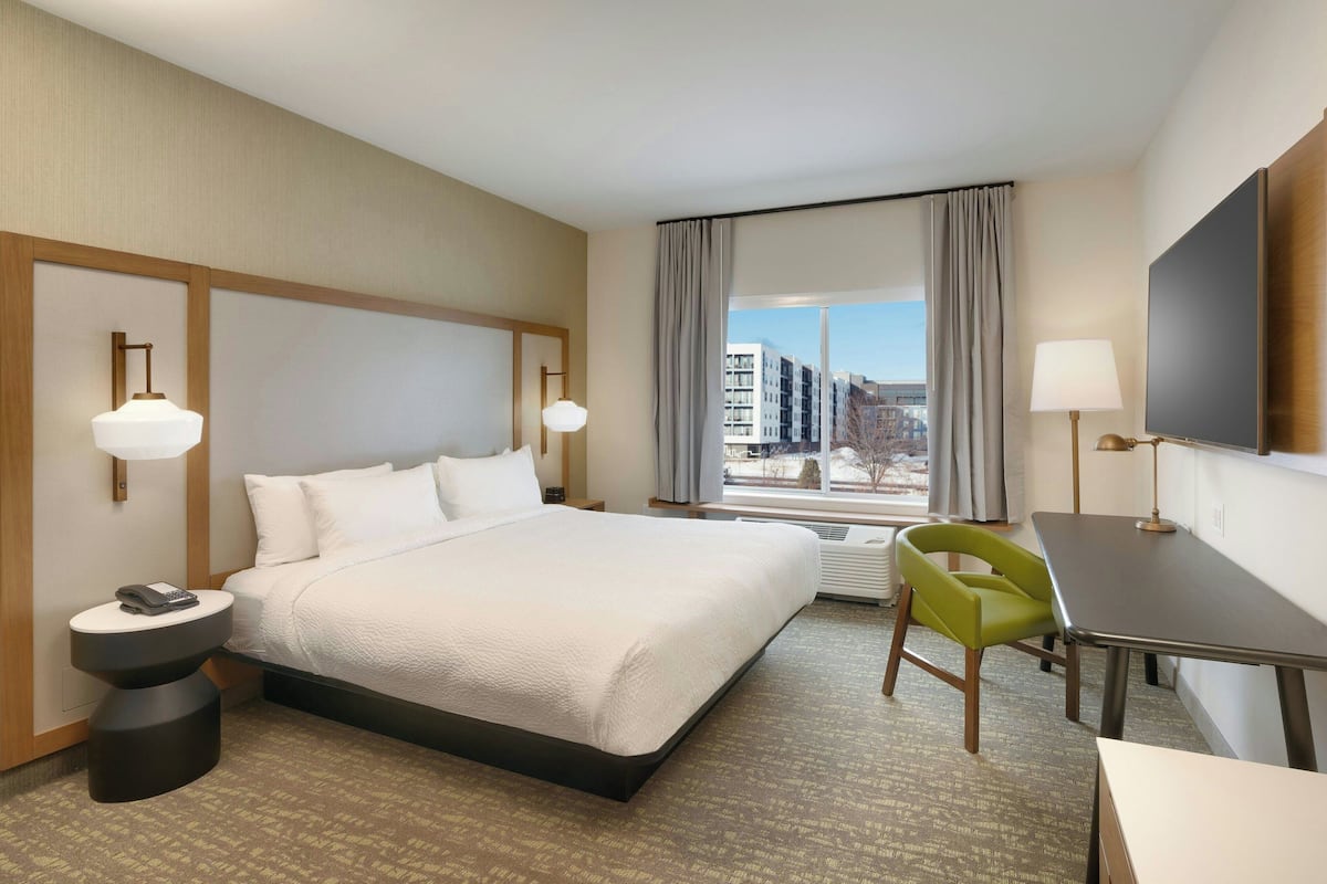 A hotel room with a large bed, a desk, a chair, a TV, and a window with a city view.