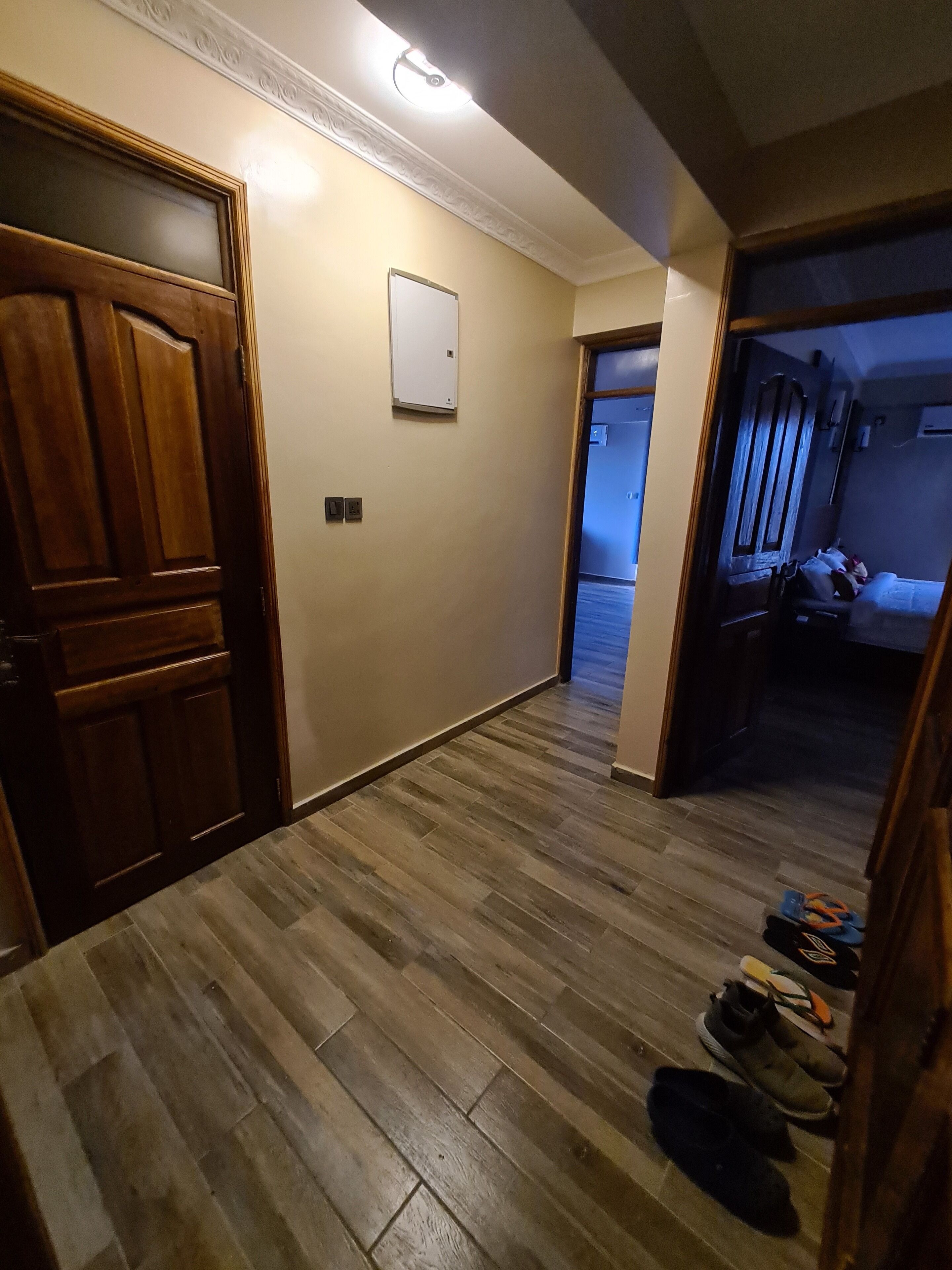 A hallway with wooden flooring, a whiteboard on the wall, and a door leading to a room with a bed and a chair.