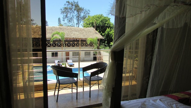 A room with a bed, a window with sheer curtains, a view of a pool and outdoor seating area.