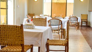 A dining area with wicker chairs and tables, light fixtures on the walls, and a view through large windows.