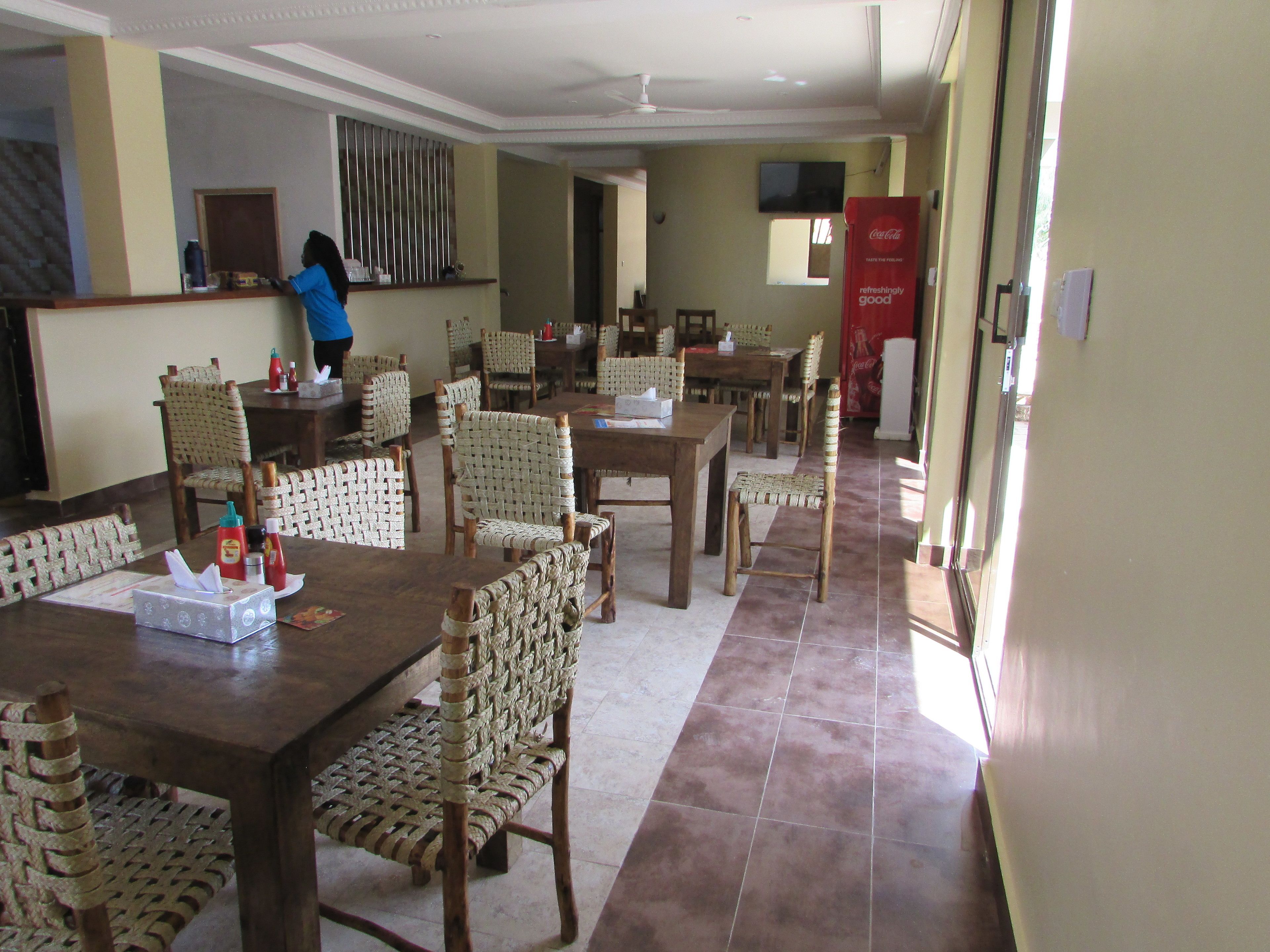 A dining area with wooden tables and woven chairs, a person in the background, and a Coca-Cola refrigerator.