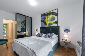 A modern bedroom with a large bed, a mirror, a decorative wall piece, and a small side table with a lamp.