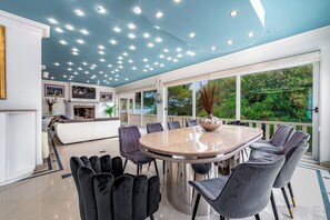 A spacious dining area with a round table and modern chairs, large windows offering a view of greenery, and a ceiling with recessed lighting.