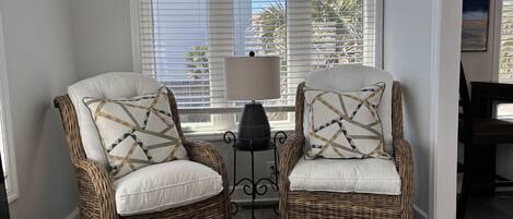 Two wicker armchairs with patterned cushions, a small table with a lamp, and a window with blinds.