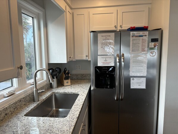 A kitchen with a stainless steel refrigerator, a granite countertop, a sink, and white cabinets. - Ocean Front Coastal Charm (Folly Beach)