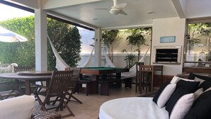 Een overdekt buitengebied met een pooltafel, rieten meubels en een zithoek met kussens.