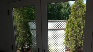 A view through a glass door leading to an outdoor deck area with wooden flooring, surrounded by potted plants and a white lattice fence. - Chatham Village - Center - Main Street Center Area (Chatham)
