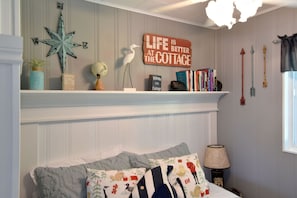 Quarto com cama, almofadas, mesa de cabeceira, abajur, estante de livros e um letreiro na parede.